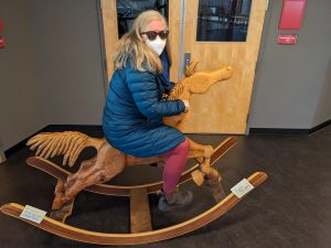 A photo of a white woman wearing sunglasses and an n95 mask on a wooden rocking horse